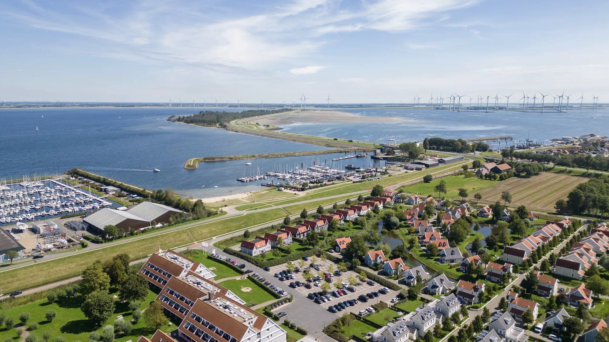 Una vista aerea di una cittadina costiera con un porto turistico, una zona residenziale e turbine eoliche in lontananza.