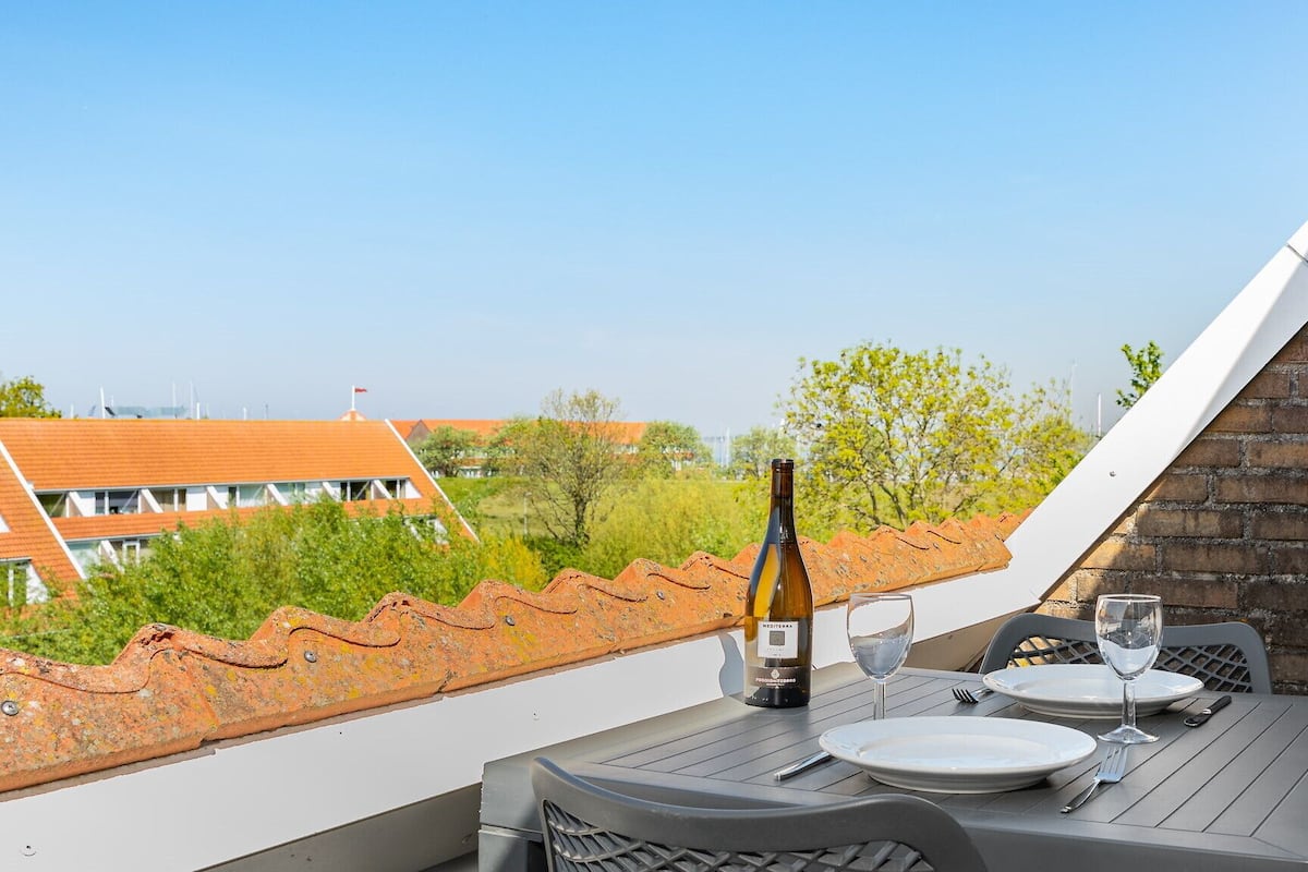 A dining table with a bottle of wine, glasses, and plates on a balcony overlooking a residential area with red-roofed houses.