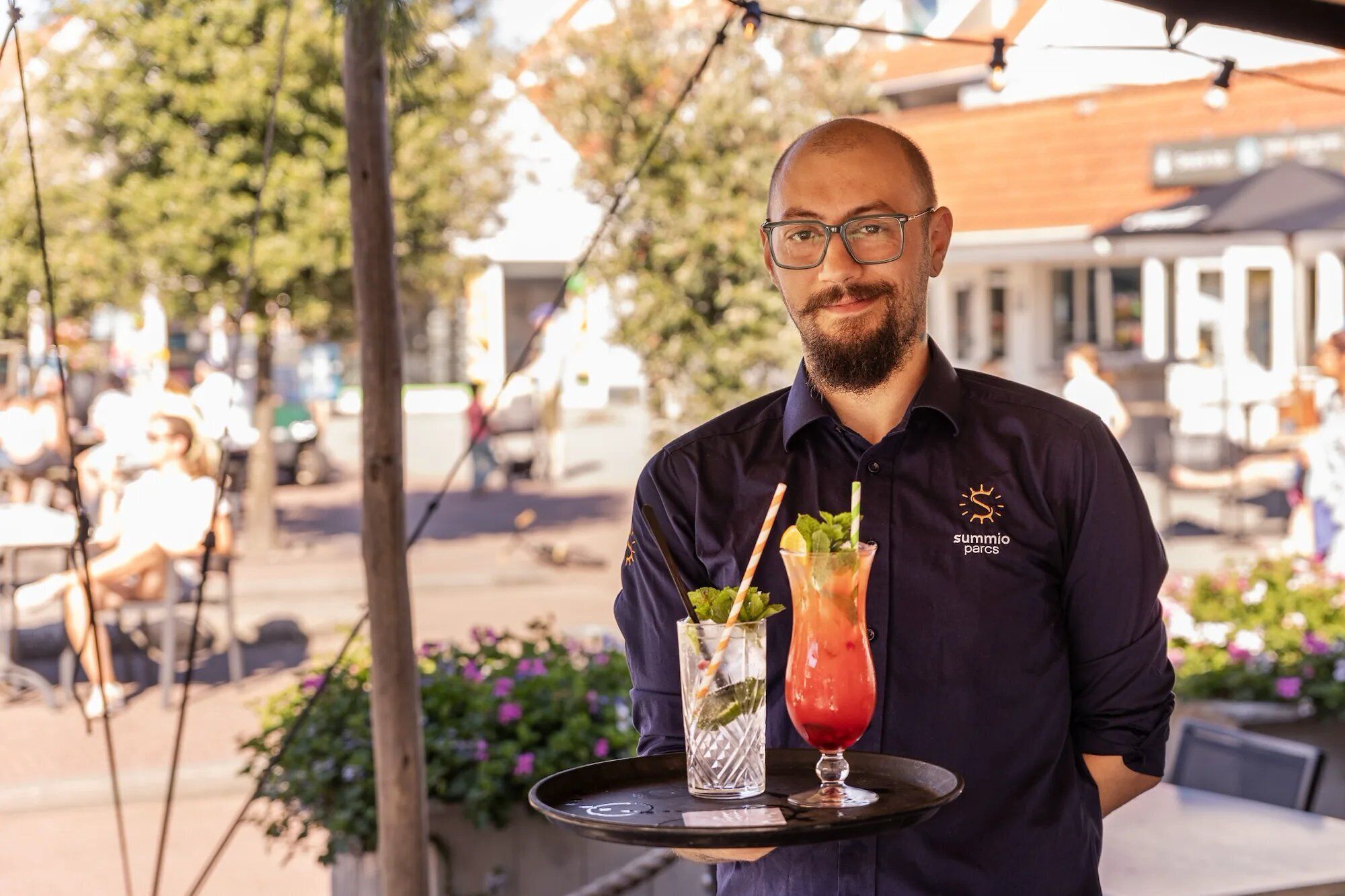 A person holding a tray with two drinks, a cocktail and a non-alcoholic beverage, in an outdoor setting with tables and chairs.