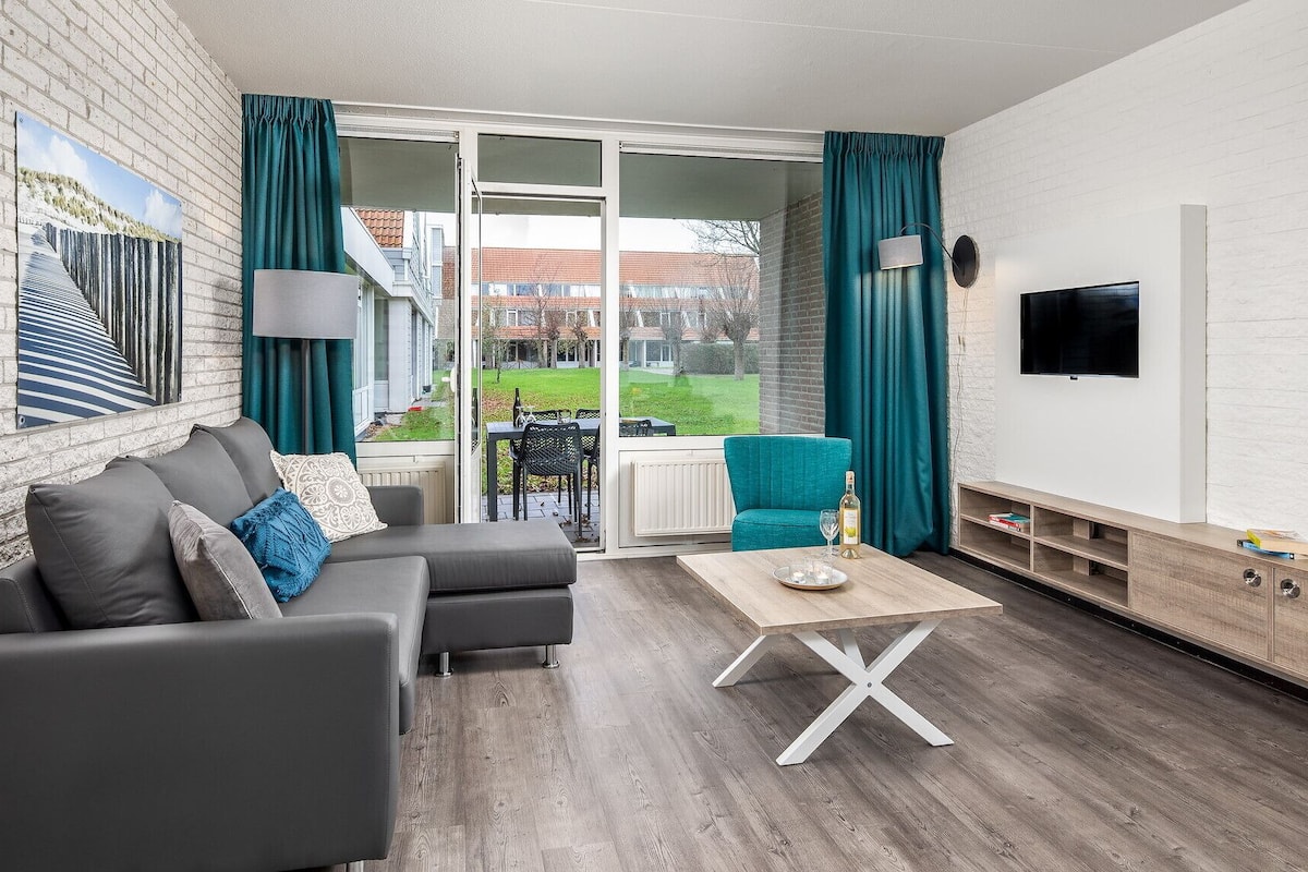 A modern living room with a grey sofa, teal curtains, a wooden coffee table, and a flat-screen TV mounted on a white brick wall.