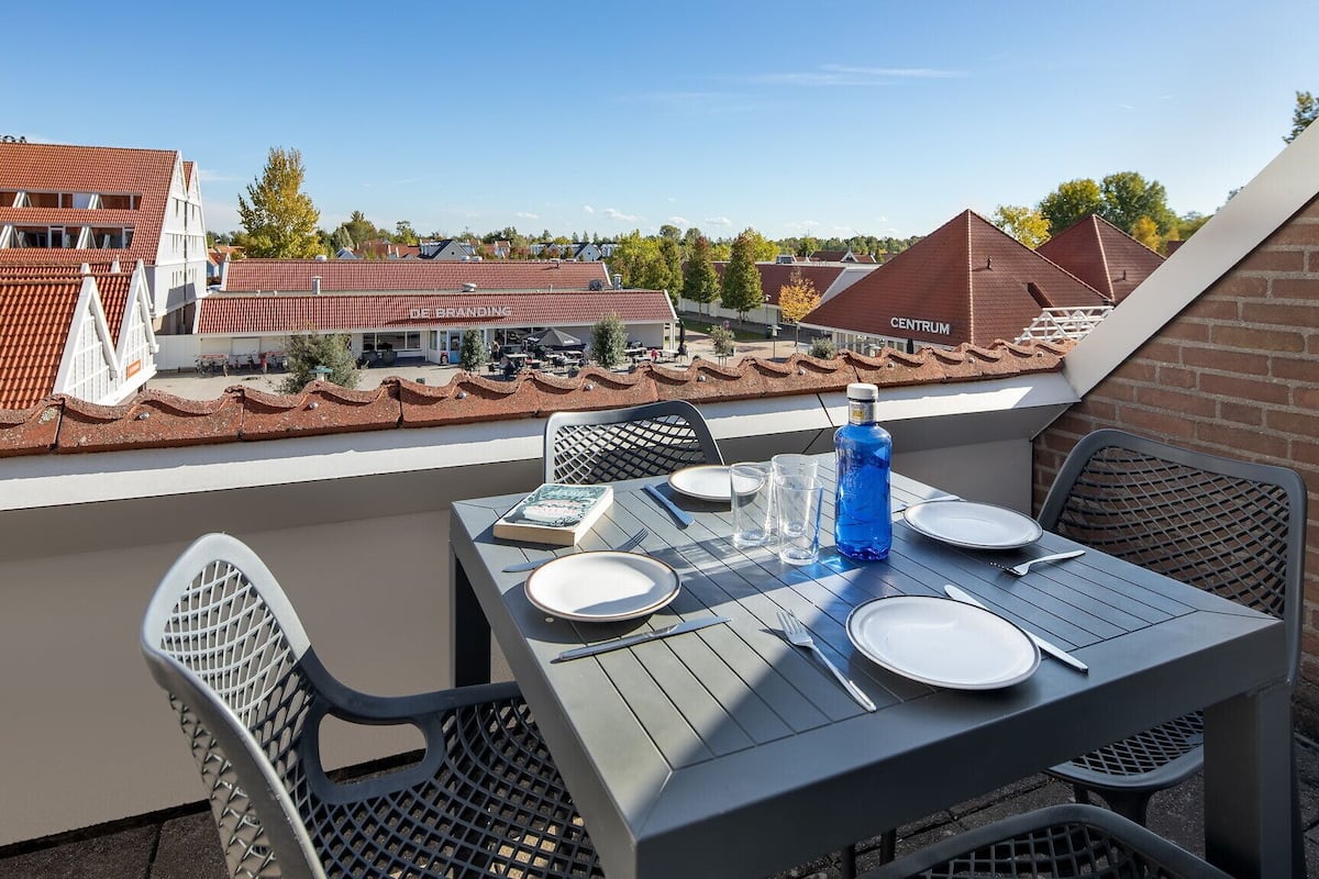 A balcony with a table set for two, overlooking a residential area with red-tiled roofs.