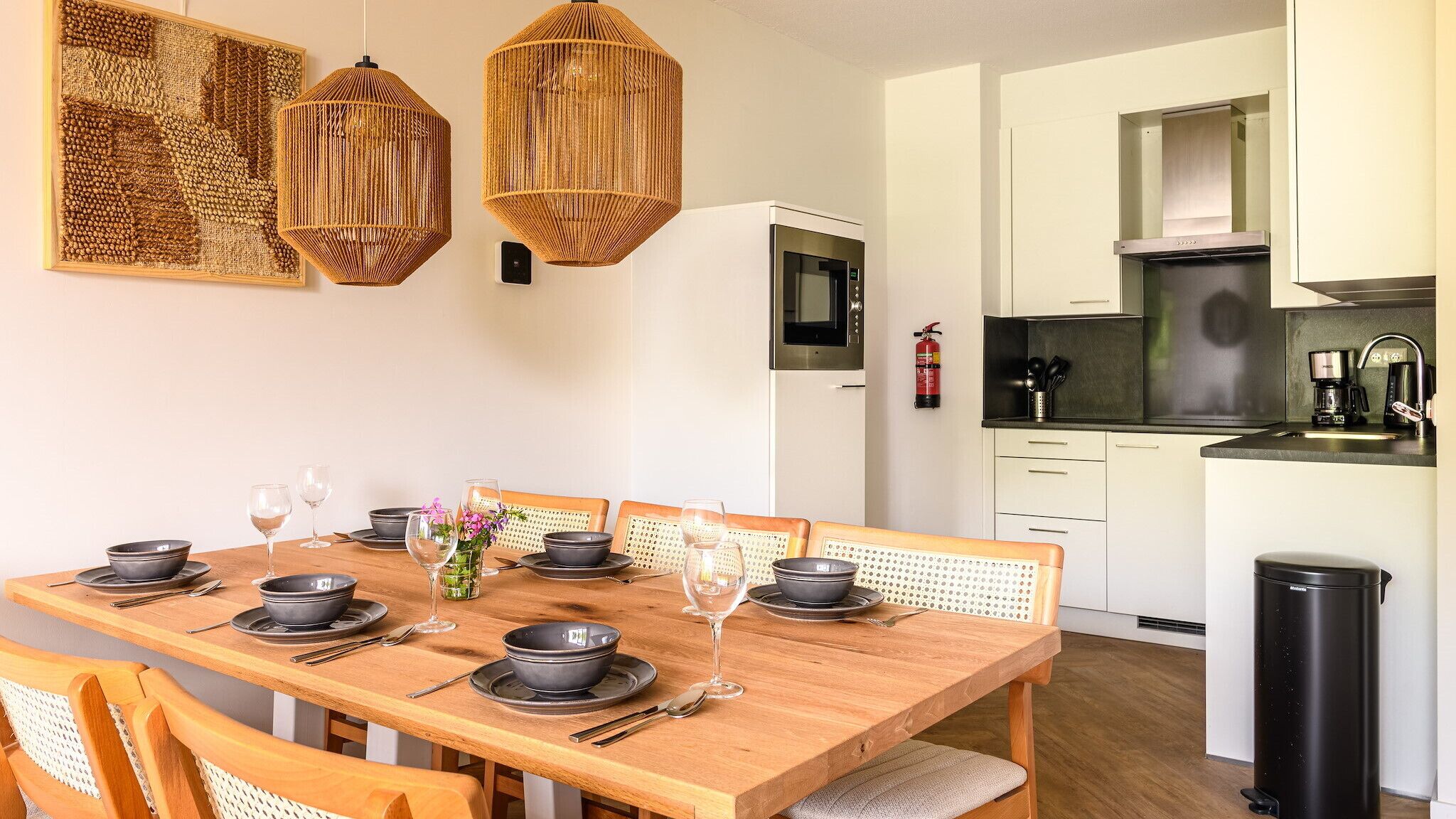 A dining area with a wooden table set for six, surrounded by wicker chairs. A kitchen with white cabinets and stainless steel appliances is visible in the background.