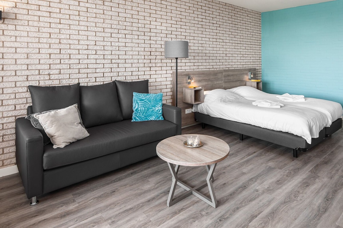 A modern hotel room with a brick wall, a grey sofa, a round wooden coffee table, and a bed with white linens.