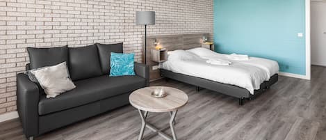 Ein modernes Hotelzimmer mit Backsteinwand, einem grauen Sofa, einem runden Holztisch und einem Bett mit weißen Laken.