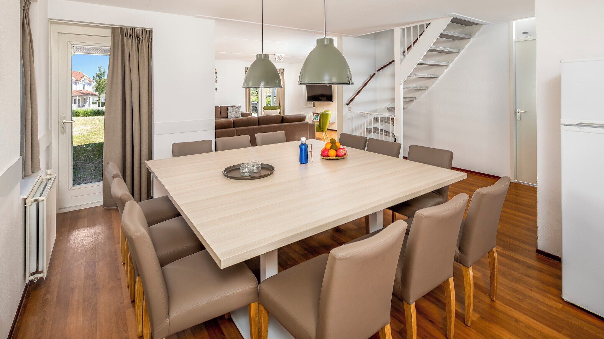 A modern dining area with a large table, multiple chairs, pendant lights, and a staircase leading to an upper level.