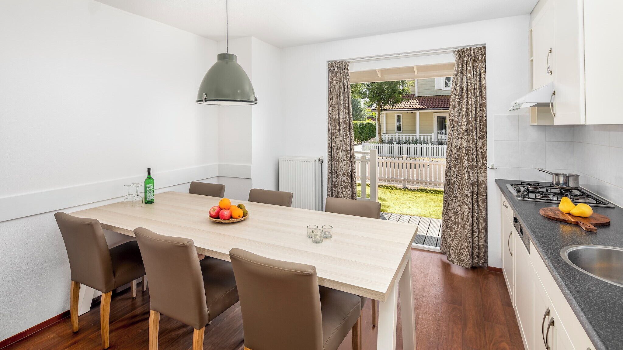 A modern kitchen with a dining area, featuring a wooden table, chairs, a hanging light, and a view of a garden.