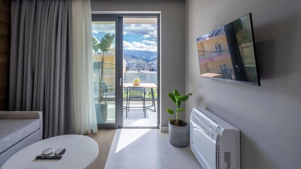 A modern living room with a sofa, a coffee table, a TV mounted on the wall, and a sliding glass door leading to a balcony with a view of buildings and mountains.