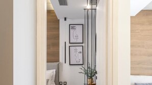 A modern hallway leading to a bedroom with a bed, bedside table, and a small seating area with a sofa and a plant.