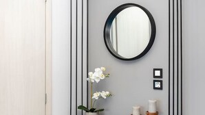 A modern bathroom with a grey wall, a black floating shelf holding a potted orchid, two candles, and a round mirror.