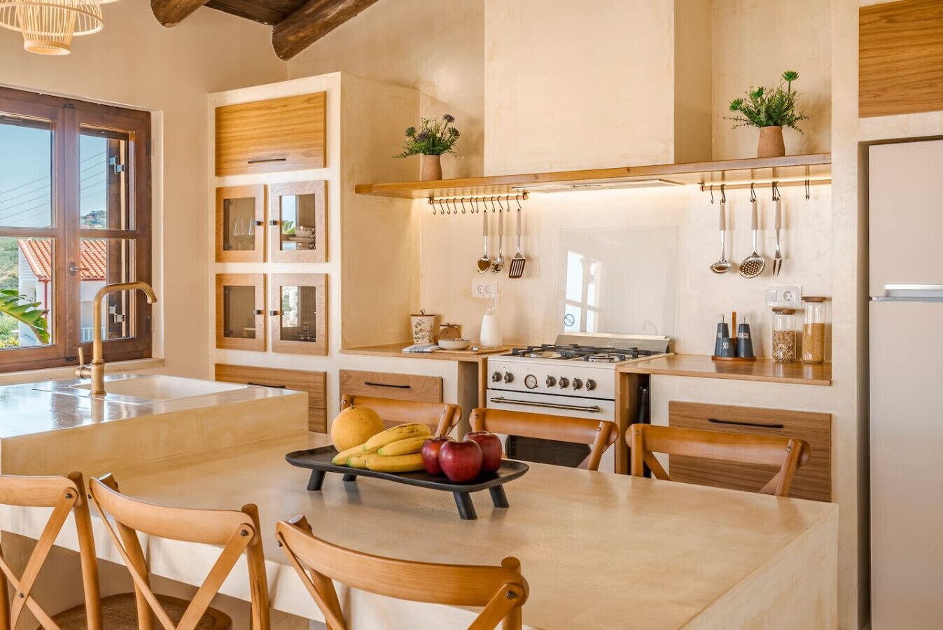 Een moderne keuken met een centraal eiland, houten kastjes en een kookplaat. Er hangen keukengerei en staat een plant in een pot aan de muur.