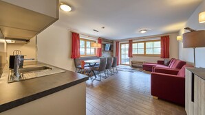 Une cuisine moderne dotée d’une cuisinière intégrée, un coin repas avec une table et des chaises, et un canapé rouge dans un salon agrémenté de grandes fenêtres.