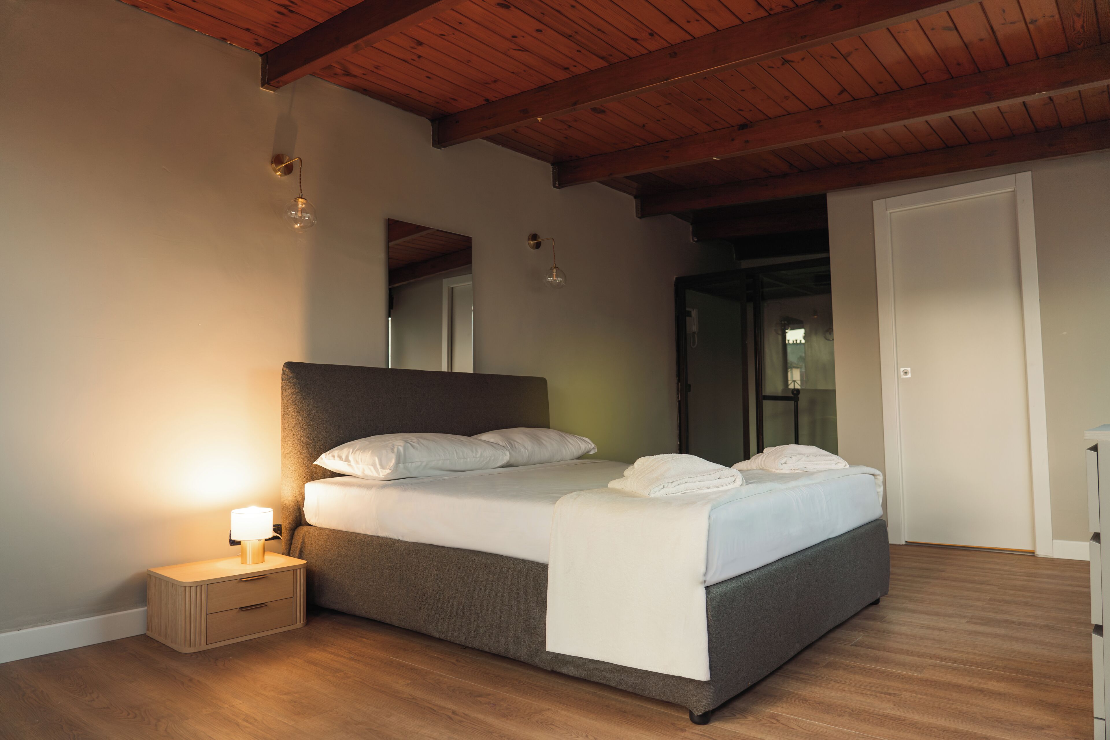 A bedroom with a wooden ceiling, a bed with white bedding, a nightstand with a lamp, and a door to another room.