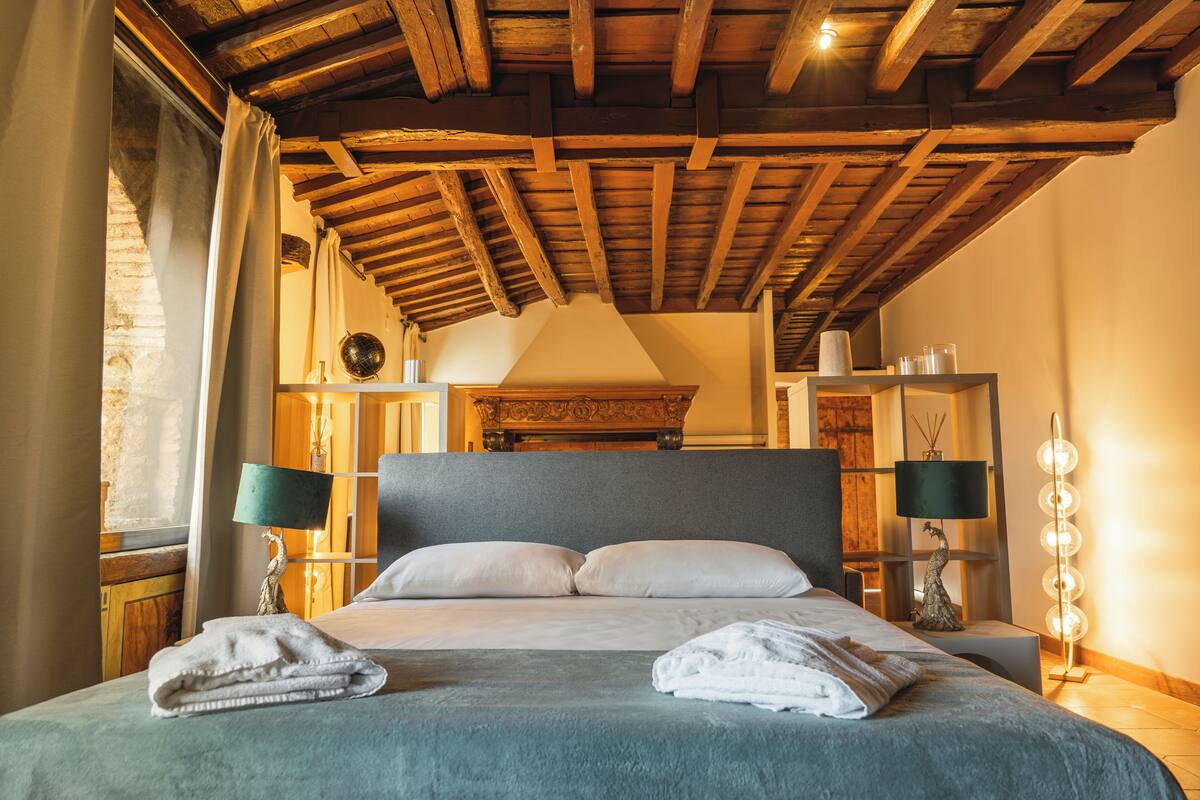 Une chambre à coucher avec un grand lit, des poutres apparentes en bois au plafond, une table de chevet avec une lampe et un luminaire fixé au mur.