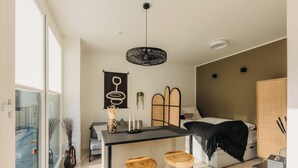 Una habitación moderna y bien iluminada con un ventanal grande, una zona de comedor con taburetes, una cama y una mesita con velas.