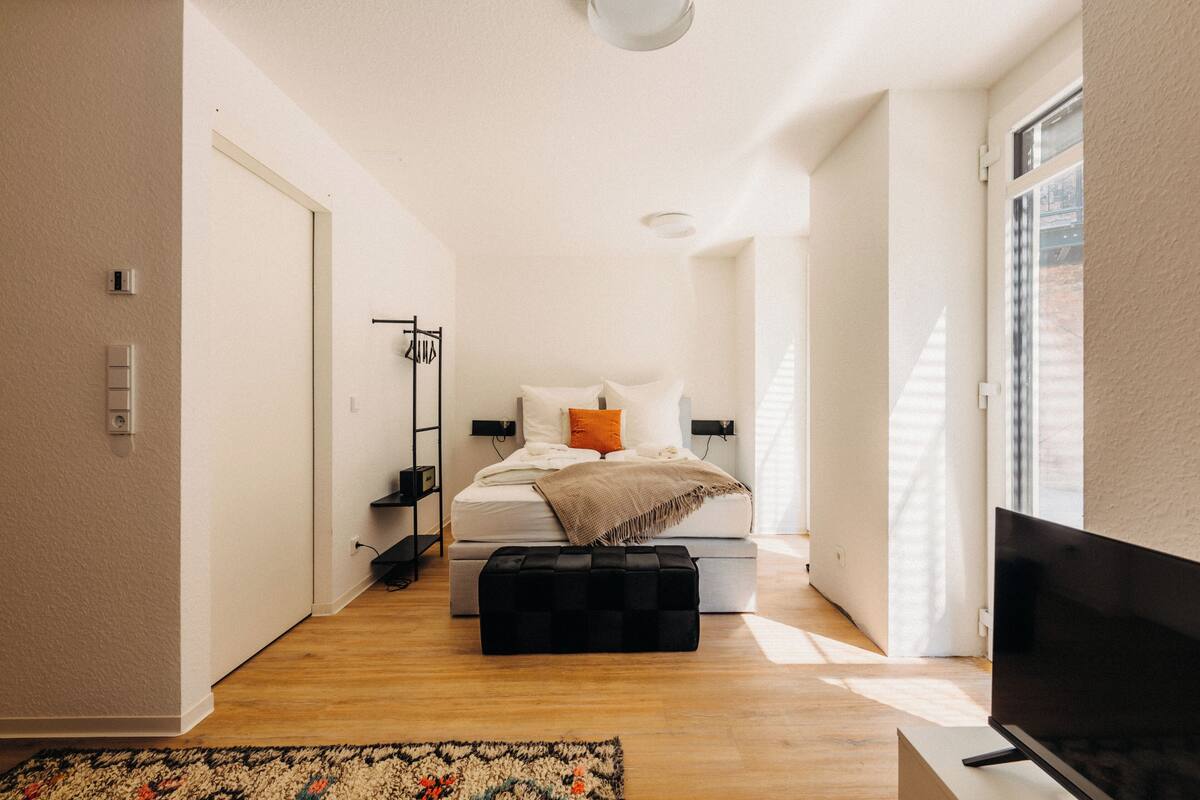 Una camera da letto moderna con un letto, un poggiapiedi nero, una TV e una finestra con persiane.