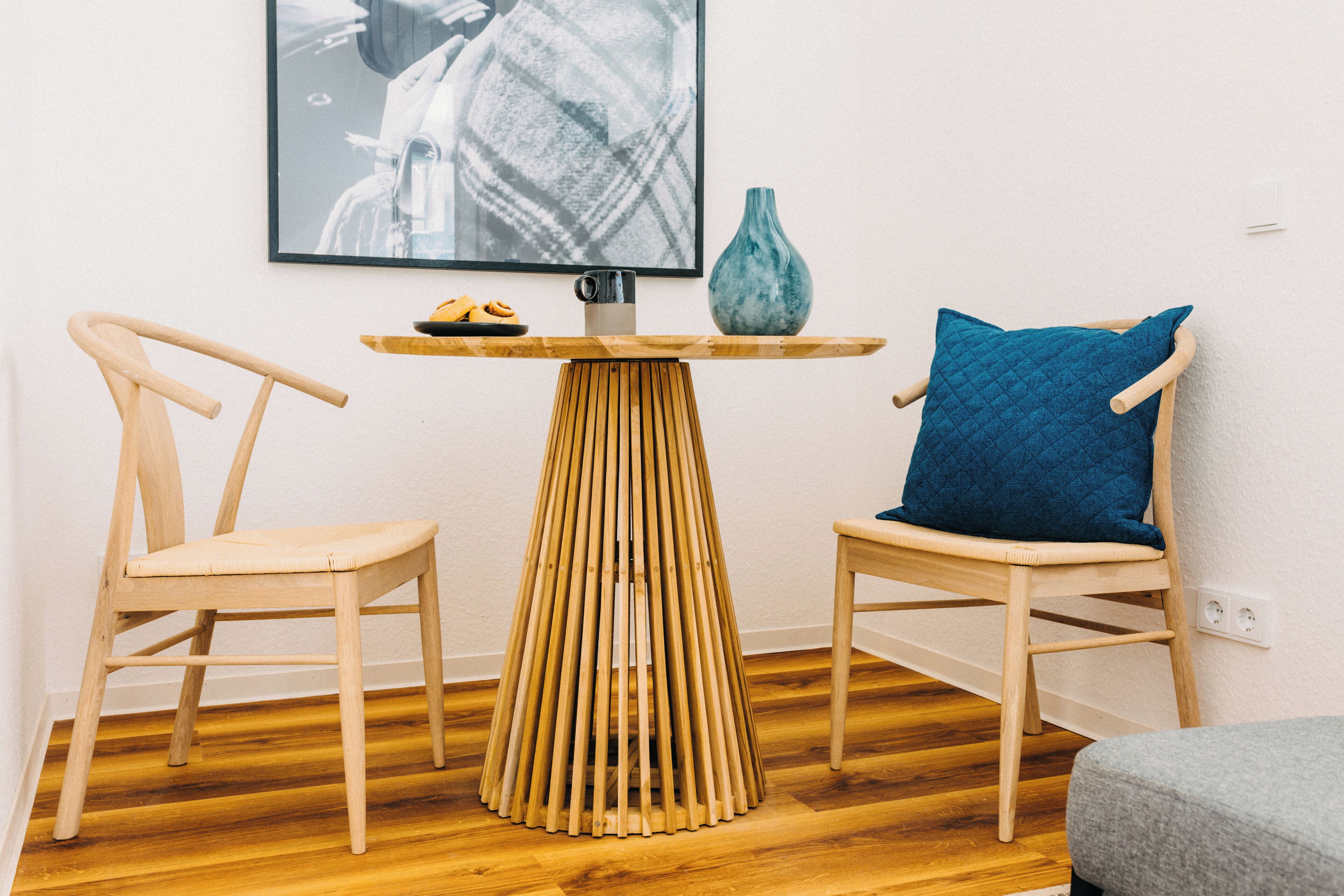 Ein Zimmer mit zwei Holzstühlen, einem kleinen Holztisch mit einer Vase und einer Schüssel, sowie einem gerahmten Bild an der Wand.