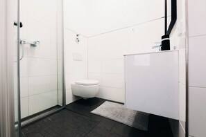A modern bathroom with a glass shower enclosure, a white toilet, and a dark floor. - Brick Lofts by BHH Living (Düsseldorf)