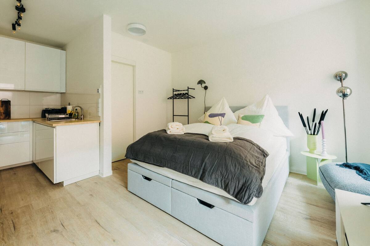 Una camera da letto moderna con un letto, un comodino e una piccola scrivania. È presente un angolo cottura con mobili e un lavandino.