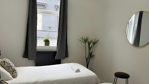A hotel room with two beds, a window with curtains, a small potted plant, a round mirror, and a stool. - Pet Friendly - Parking - 3 Bedrooms - 6 Guests (Canterbury)