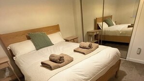 A single bed with a wooden headboard, white bedding, and two rolled towels. A small bedside table and a mirror are visible. - Heathercroft Apartment-Modern Comfort in Fort William. (Fort William)