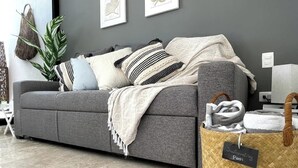 Un canapé gris agrémenté de plusieurs coussins et d’une couverture, un panier tressé avec une étiquette, et une plante dans une pièce aux murs gris et au sol carrelé.