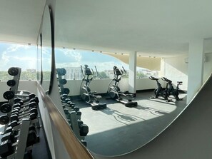 Une salle de sport équipée de matériel d’exercice, notamment des haltères, des tapis de course et des vélos d’appartement, et offrant une vue sur la ville grâce à de grandes fenêtres.