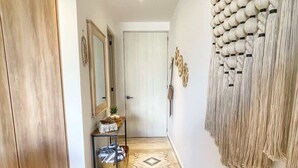 Un couloir avec une porte en bois, un miroir, une petite table sur laquelle se trouve un panier, et une décoration murale en macramé.