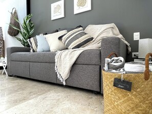 Un salon moderne avec un canapé gris, des coussins à motifs, un panier tressé muni d’une étiquette et une plante en pot.