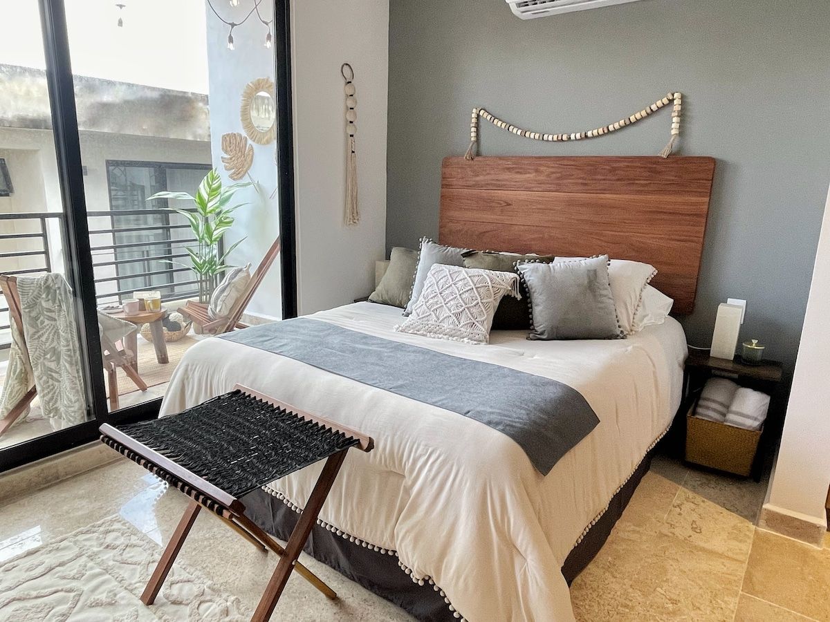A bedroom with a bed, a small table, a chair, a bedside table, and a wall-mounted air conditioner.