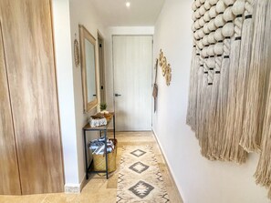 Um corredor com porta de madeira, espelho, mesa pequena com cesta, tapete estampado e um objeto pendurado na parede.