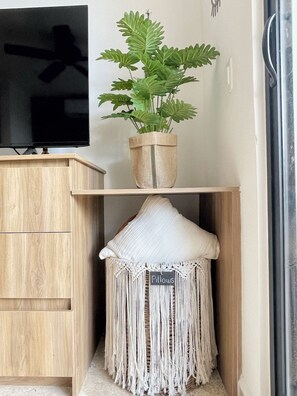 A wooden cabinet with a television on top, a potted plant, and a macramé pillow labeled 'Pillows' on a shelf.