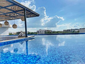 Une piscine avec un bord carrelé, un espace de repos couvert avec des chaises, et un ciel dégagé.