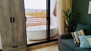 Un salon moderne avec un canapé gris, une grande plante en pot et une vue sur un balcon où se trouve une baignoire blanche.