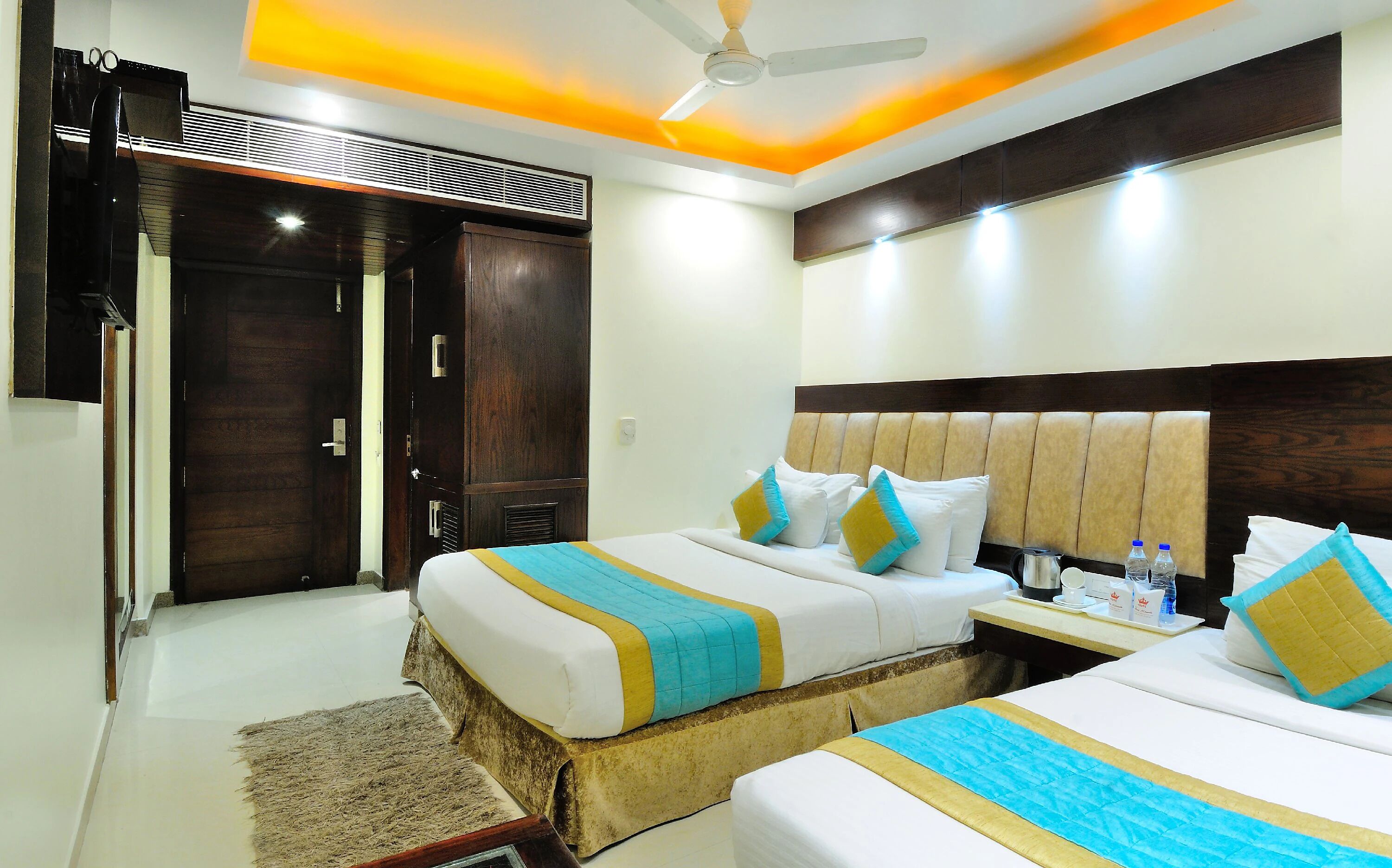 A hotel room with two beds, a ceiling fan, and a nightstand with a kettle and water bottles.