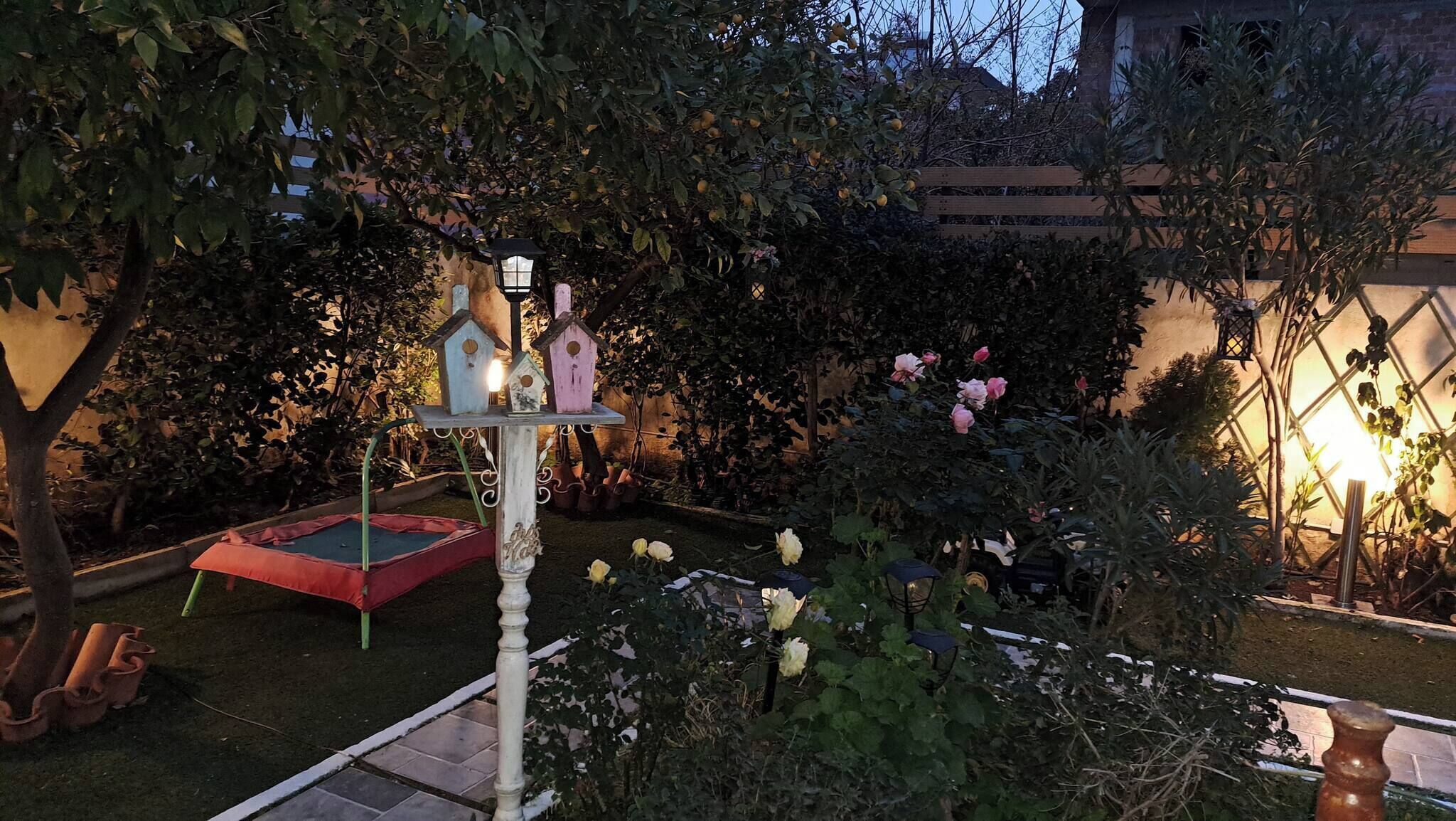 Un giardino con una struttura decorativa a forma di casetta per uccelli, una piccola area giochi rossa e fiori in fiore.