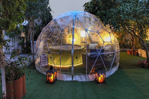 A transparent geodesic dome structure with a central seating area, surrounded by artificial grass and potted plants.