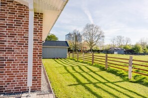 Un bâtiment en briques surmonté d’une marquise blanche, une zone délimitée par une clôture avec de l’herbe, et un petit abri de jardin à l’arrière-plan.