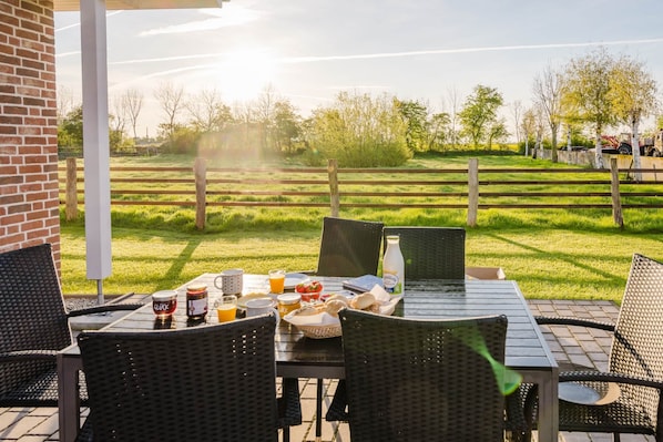 An outdoor patio with a table set for breakfast, including bread, butter, and juice, overlooking a grassy field and trees.