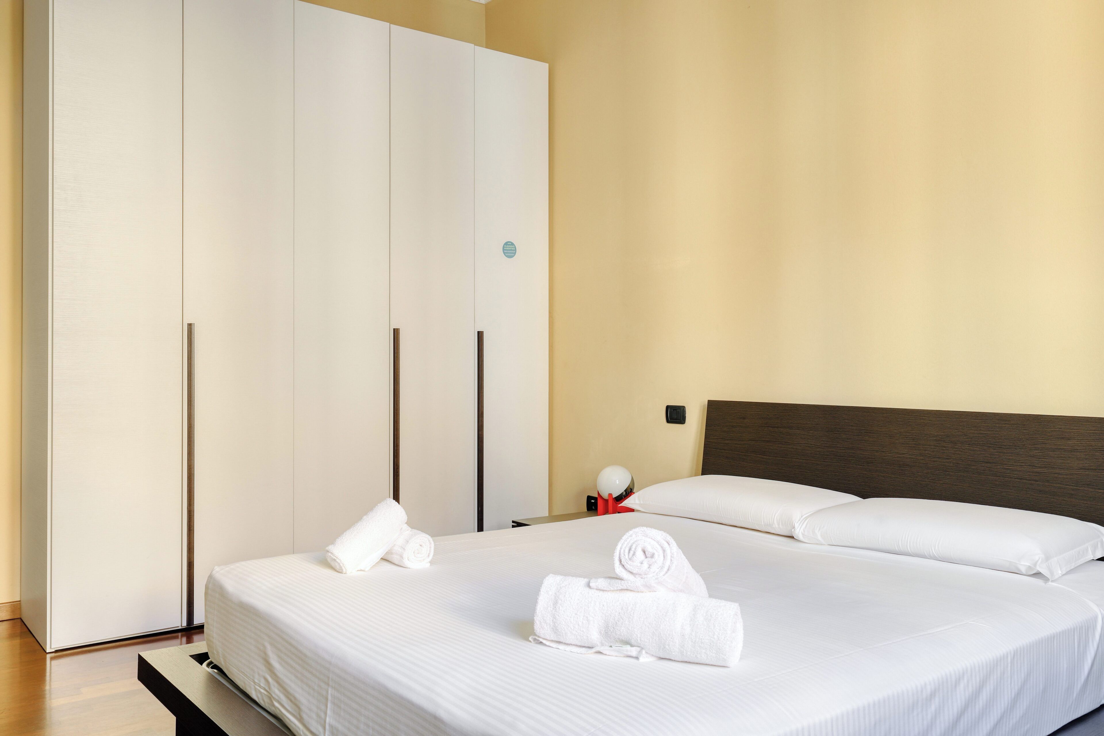 A hotel room with a large bed, a wardrobe, and a bedside table with a lamp.