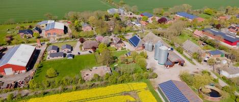 Eine Vogelperspektive auf eine ländliche Gegend mit Windturbinen, Feldern voller gelber Blumen und verschiedenen Gebäuden, darunter ein großes Getreidespeicher und Solaranlagen.