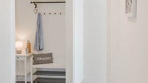 Un pasillo con una puerta blanca, un perchero con una chaqueta, una mesita con una lámpara, un cojín con texto y una planta en maceta.