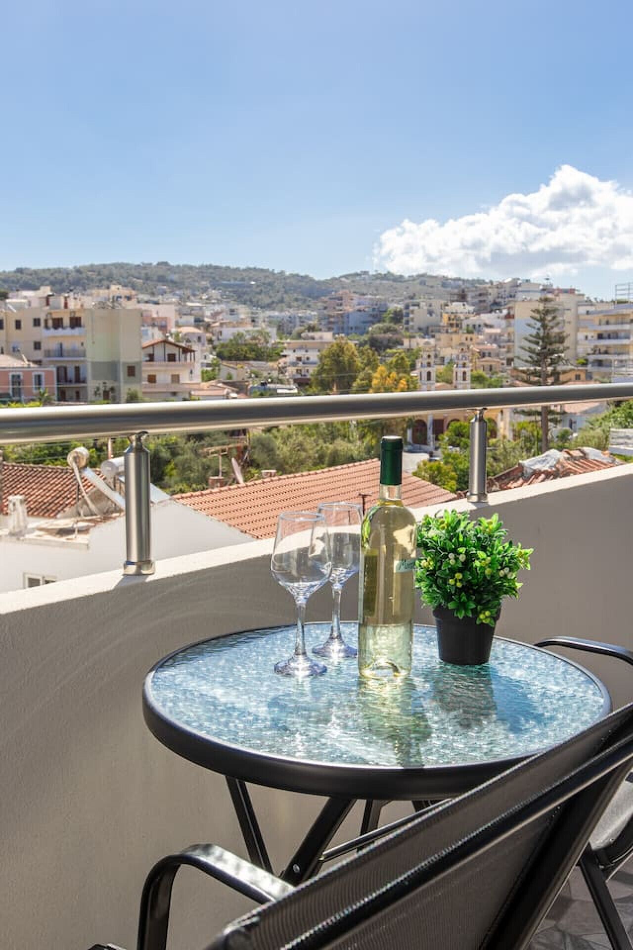 Une table ronde en verre, sur laquelle se trouvent une bouteille de vin blanc, deux verres à vin et une plante en pot, est placée sur un balcon donnant sur un paysage urbain avec des bâtiments et des arbres.