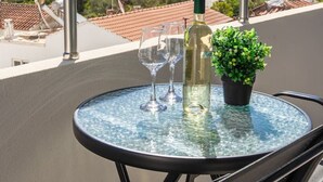 Een ronde glazen tafel met een fles witte wijn, twee wijnglazen en een potplant op een balkon met uitzicht op een stadsgezicht met gebouwen en bomen.