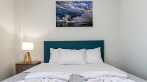 Een keurig opgemaakt bed met witte lakens en handdoeken, een blauwgroen hoofdbord en een schilderij van een vuurtoren aan de muur.