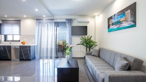 Een moderne woonkamer met een grijze bank, een zwarte salontafel en een aan de muur bevestigde televisie. Er is een keuken met een spoelbak en kasten, en een groot raam met gordijnen.