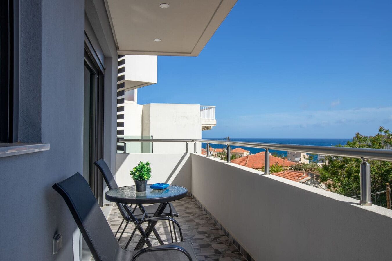 A balcony with a table and chairs, a potted plant, and a view of the sea and buildings.