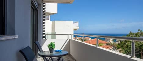 Un balcón con una mesa y sillas, una planta en maceta y vistas al mar y edificios.