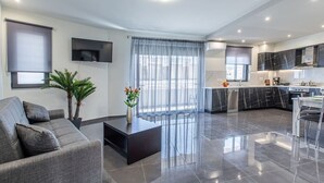 Een moderne woonkamer met een grijze bank, een salontafel en een televisie. Er is een keuken met zwarte kasten en een eethoek met een tafel en stoelen.