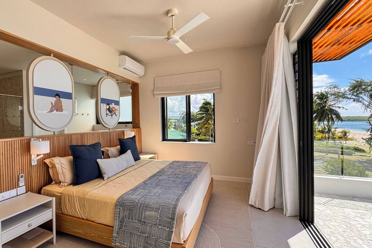 Un dormitorio con cama, mesita de noche, espejo, ventilador de techo y ventana con vista a la playa.