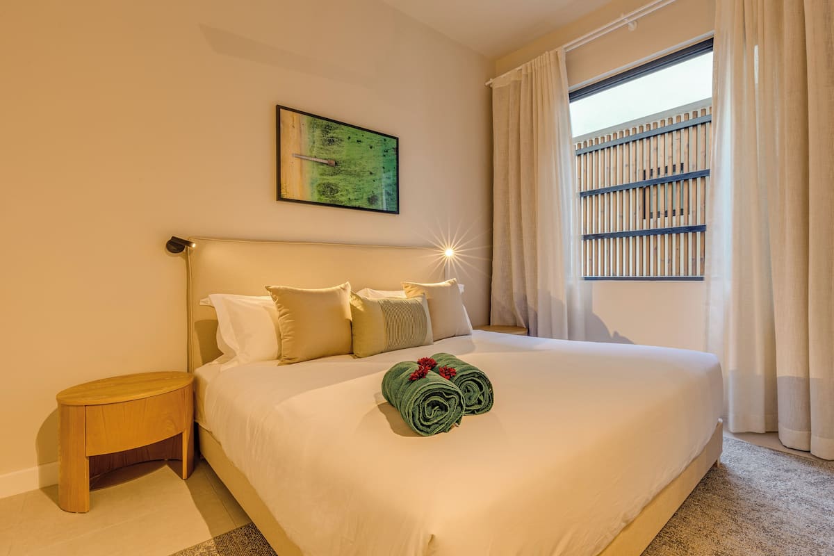 Una habitación de hotel con una cama bien hecha, una mesita de noche de madera, un cuadro en la pared y una ventana con cortinas.